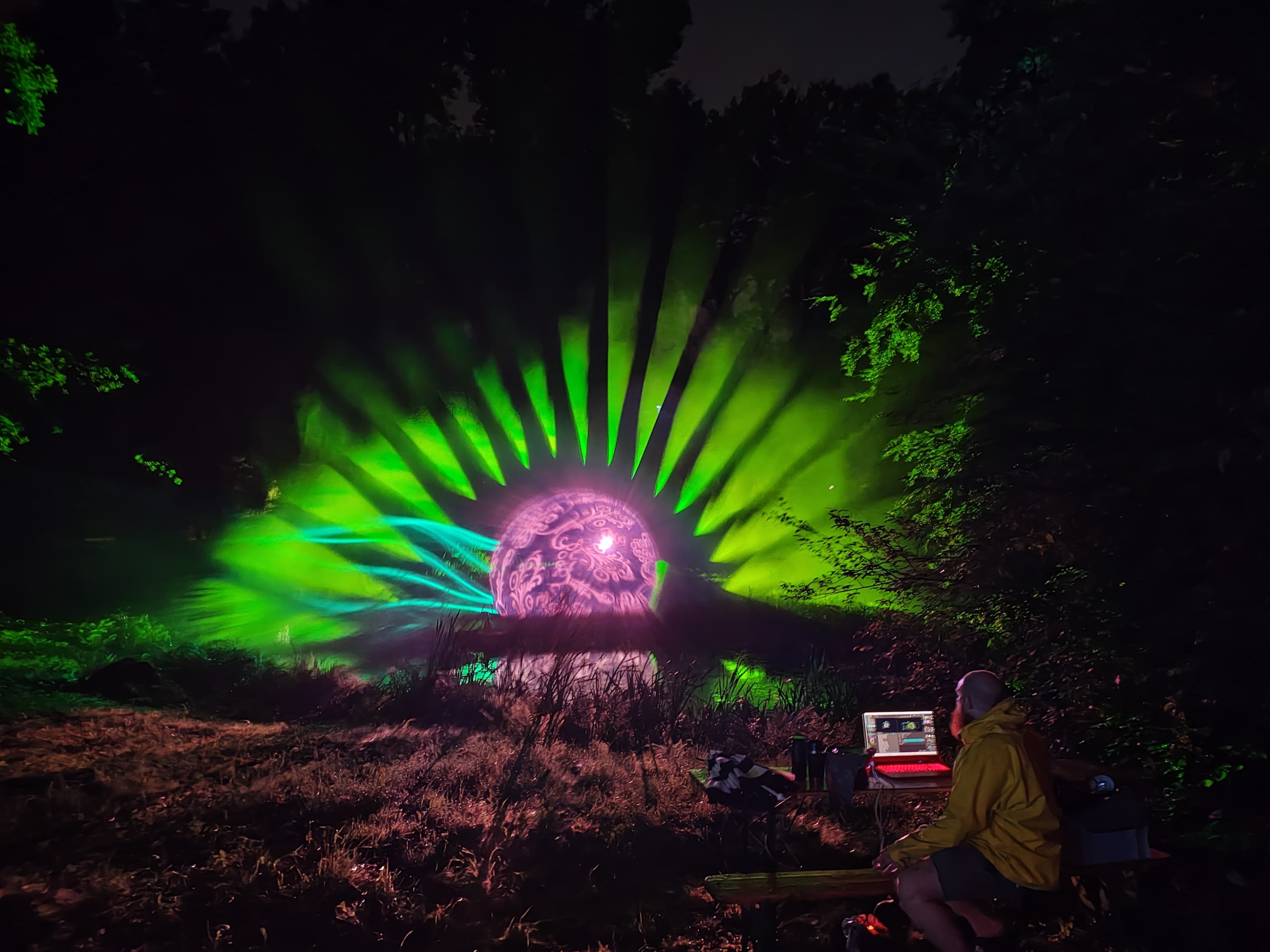 Aufnahme beim Park- & Blütenfest in Kromlau 2025; Foto: Daniel Häfner Magentafarbene Lichtkugel auf einer Wiese im Kromlauer Park zwischen Sträuchern und Bäumen, von der Kugel gehen grüne Lichtstrahlen aus, ähnlich wie Sonnenstrahlen. Im Vordergrund sitzt ein Mann mit einem Laptop und weiterer Technik auf der Wiese und steuert die Installation.
